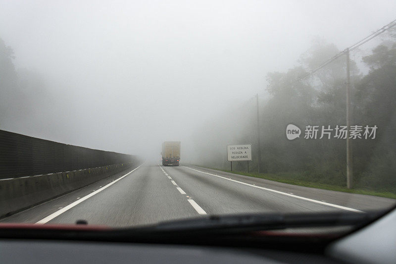 一辆卡车在有雾的道路上行驶，经过一个牌子上写着“UNDER fog reducing THE SPEED”的卡车